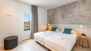 Een hotelkamer met een groot bed, een klein krukje, een kamerplant en uitzicht op buiten.