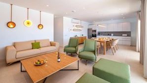 Een moderne woonkamer met een bank, groene fauteuils en een houten salontafel met fruit en wijnglazen. In de achtergrond is een keuken met een eethoek te zien.