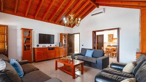 Un vaste salon doté d’un plafond en bois, d’un téléviseur à écran plat, d’un canapé, d’une table basse et d’un coin repas visible à travers une porte ouverte.