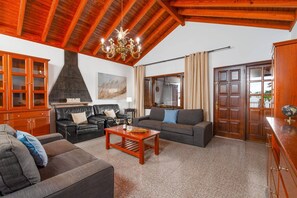 A living room with a high wooden ceiling, a chandelier, a fireplace, and a large window with curtains. - Villa Victoria Lanzarote by Villa Plus (Playa Blanca)