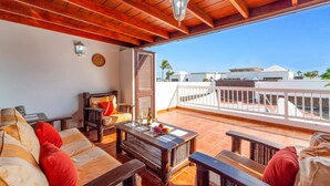 Un balcon avec des meubles en bois, une table avec une bouteille et des fruits, et une vue sur des immeubles et des palmiers.