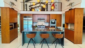 Een moderne keuken met een centraal eiland, houten kastjes, roestvrijstalen apparatuur en een entresol met een balustrade en hangende lampen.
