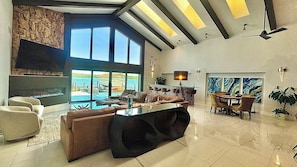 A spacious living area with a large window overlooking a pool, a fireplace, and a dining area with a view of the sea. - The Grand Bay House in Fort Myers Beach (Fort Myers Beach)