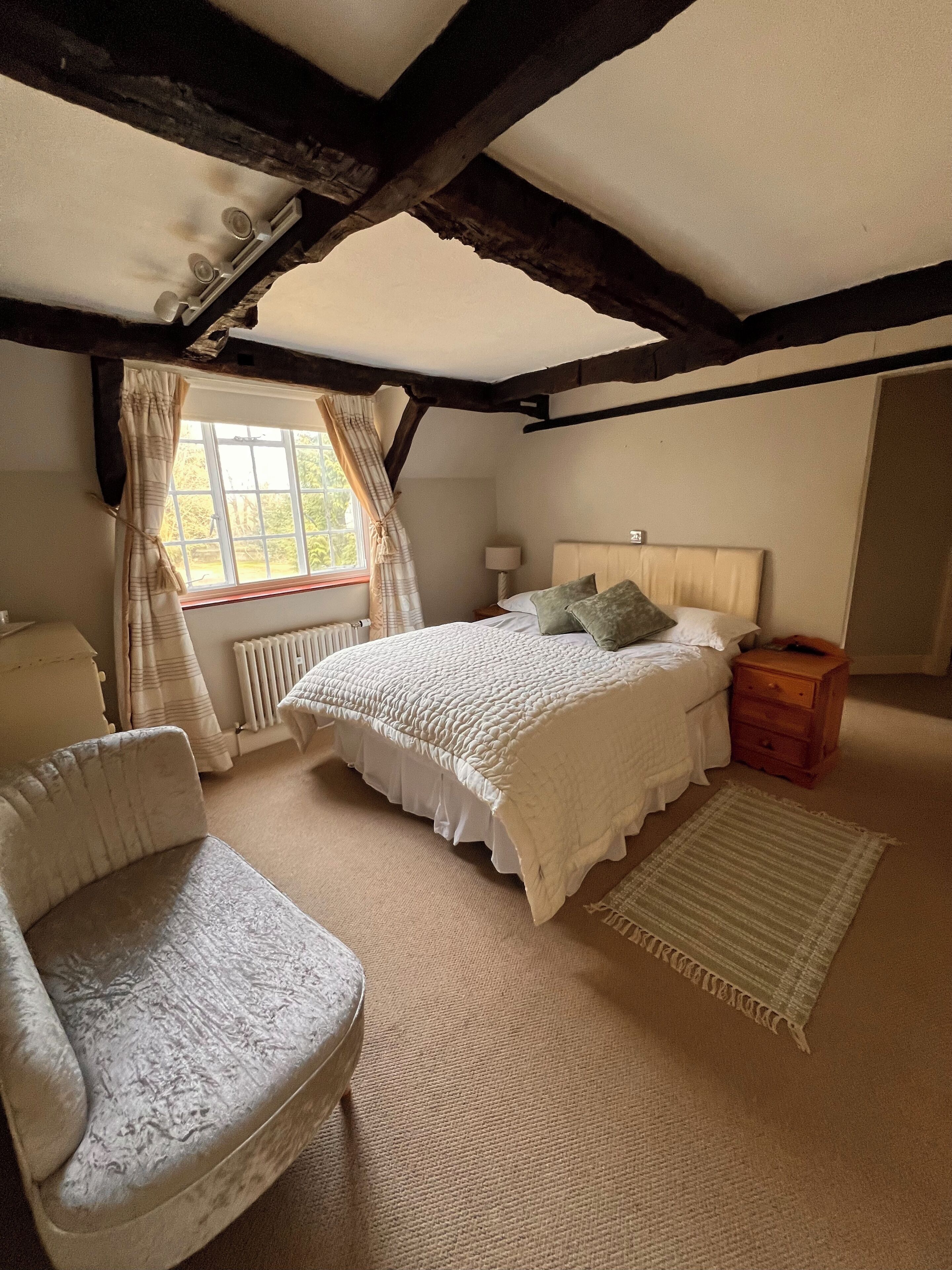 A bedroom with a bed, a chair, a nightstand, a window with curtains, and a radiator.