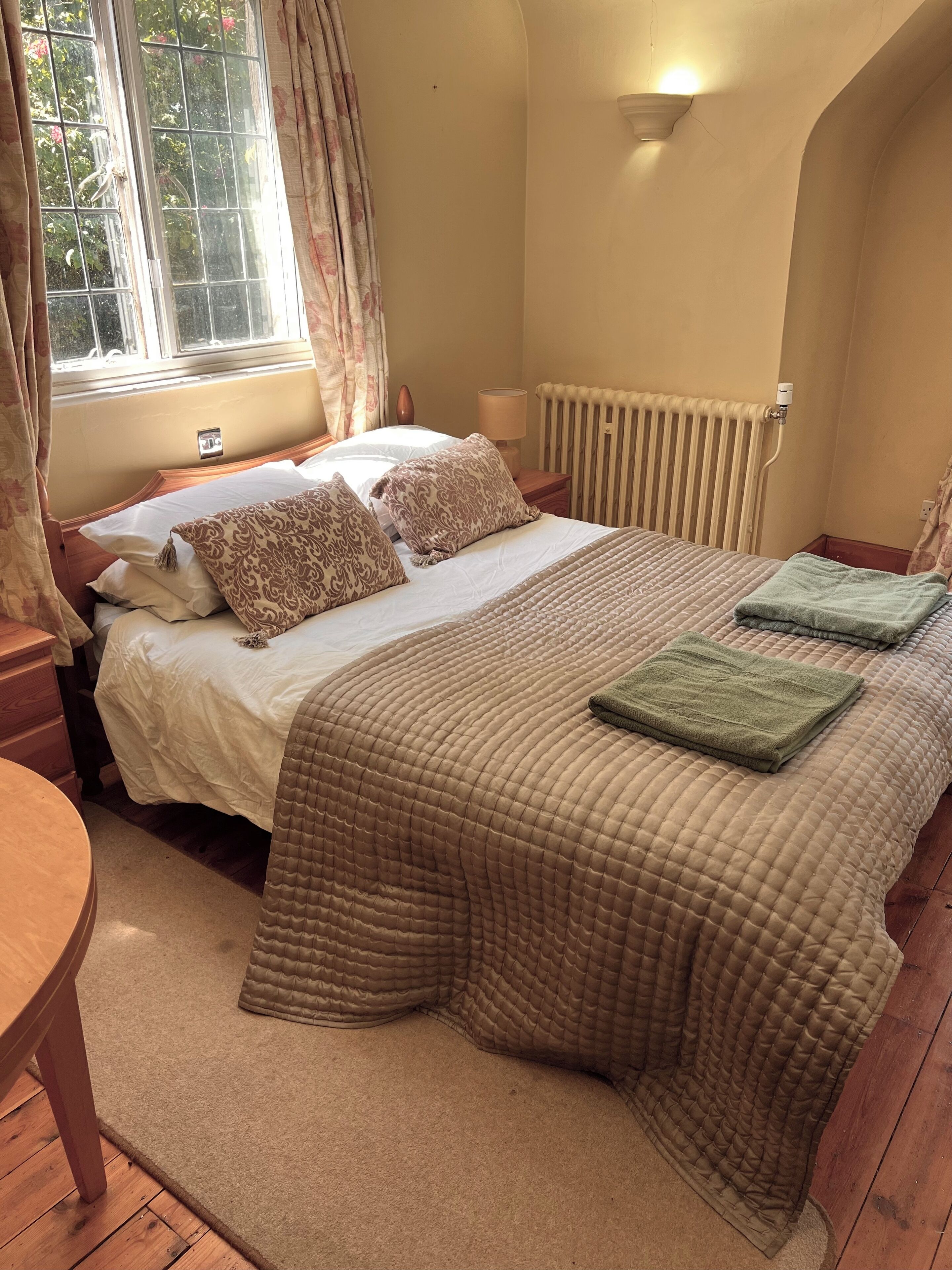 A bedroom with a bed, bedside table, lamp, window with curtains, radiator, and a view of greenery outside.