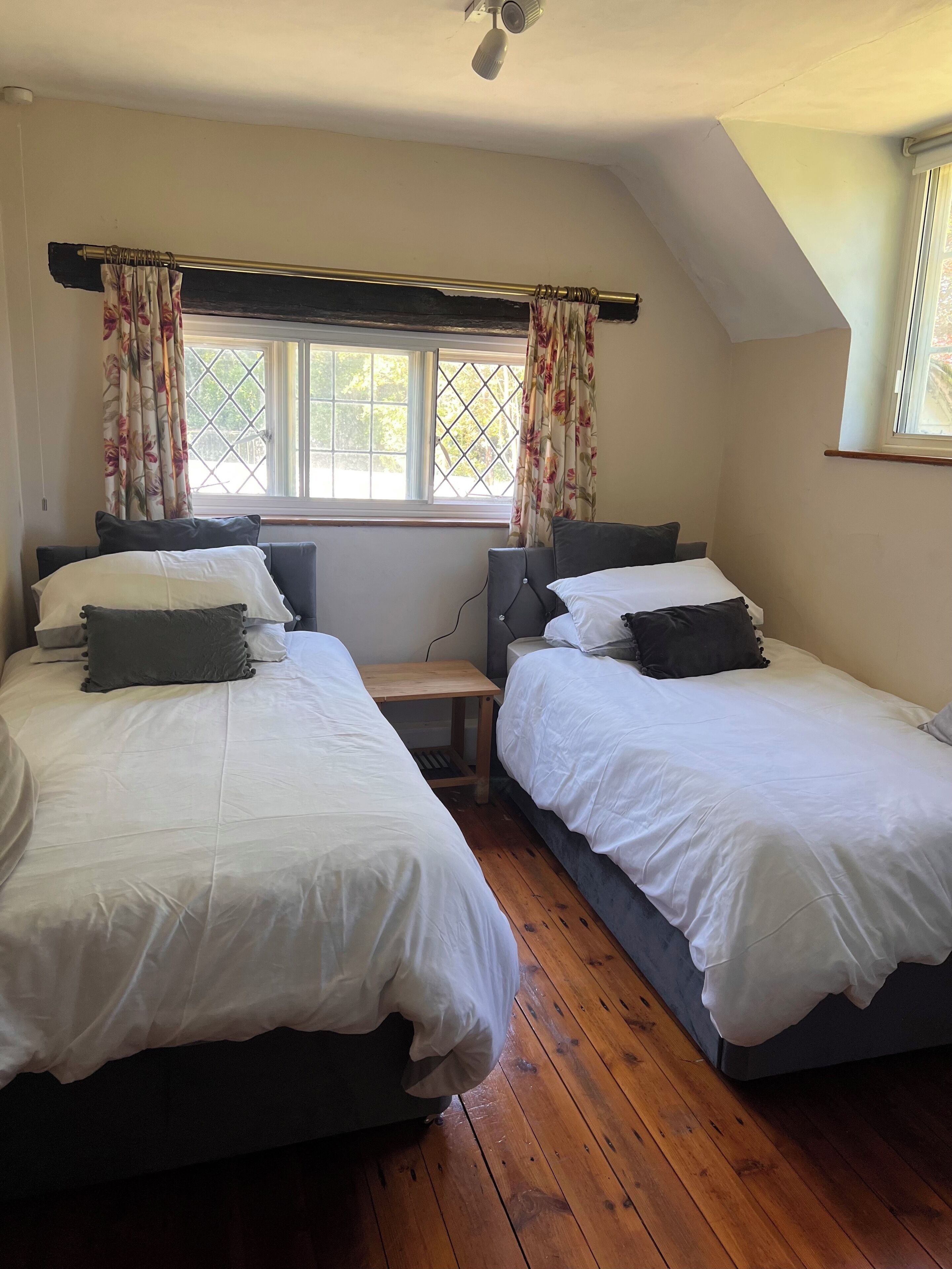 Una habitació amb dos llits individuals, sòcols de fusta, una finestra amb cortines florals i un ventilador de sostre.