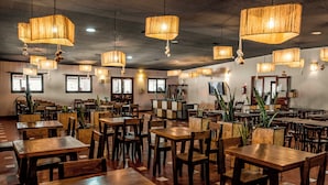 En rymlig restaurang med träbord och stolar, hängande lampor och krukväxter.