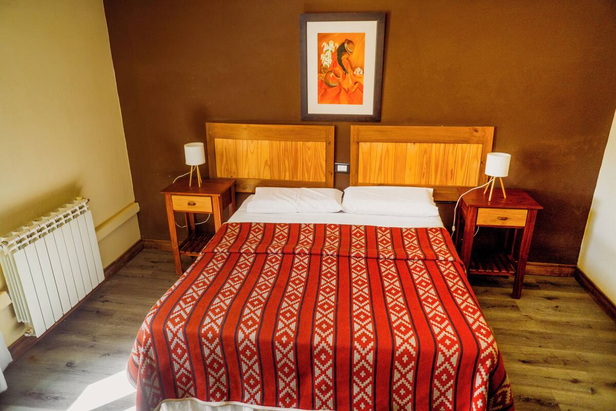 A double bed with a patterned bedspread, two bedside tables with lamps, a framed picture on the wall, and a radiator on the floor.