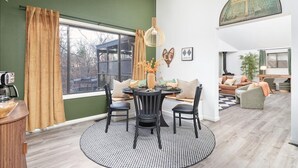 Una sala moderna con una mesa redonda de comedor, sillas negras, una mesa de centro y un sofá. Hay un ventanal con cortinas, una lámpara colgante y un cuadro en la pared.