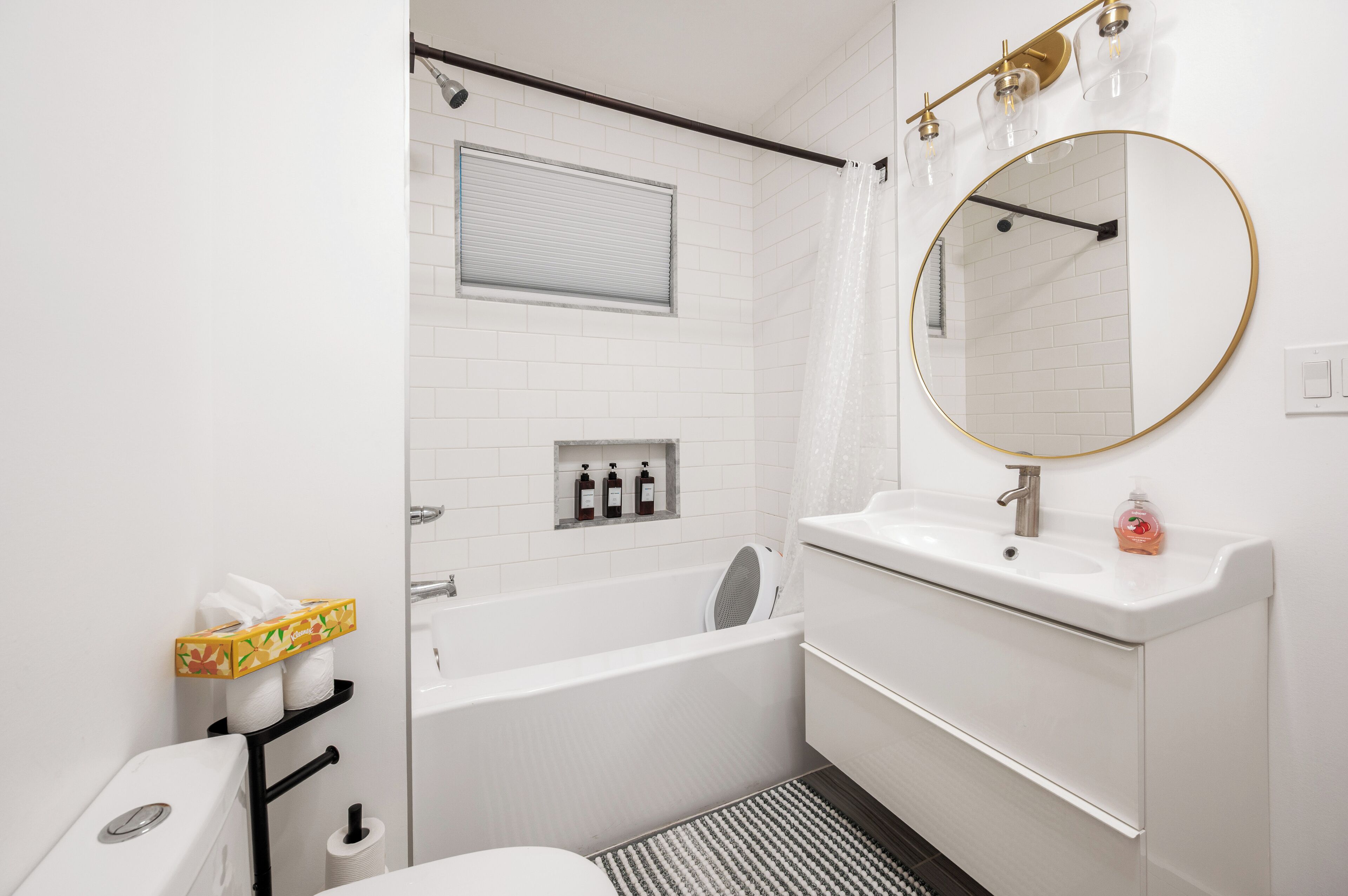 Et moderne badeværelse med en hvid vask, badekar og toilet. Der er et spejl med gylden ramme, et håndklædestativ og et vindue med persienner.