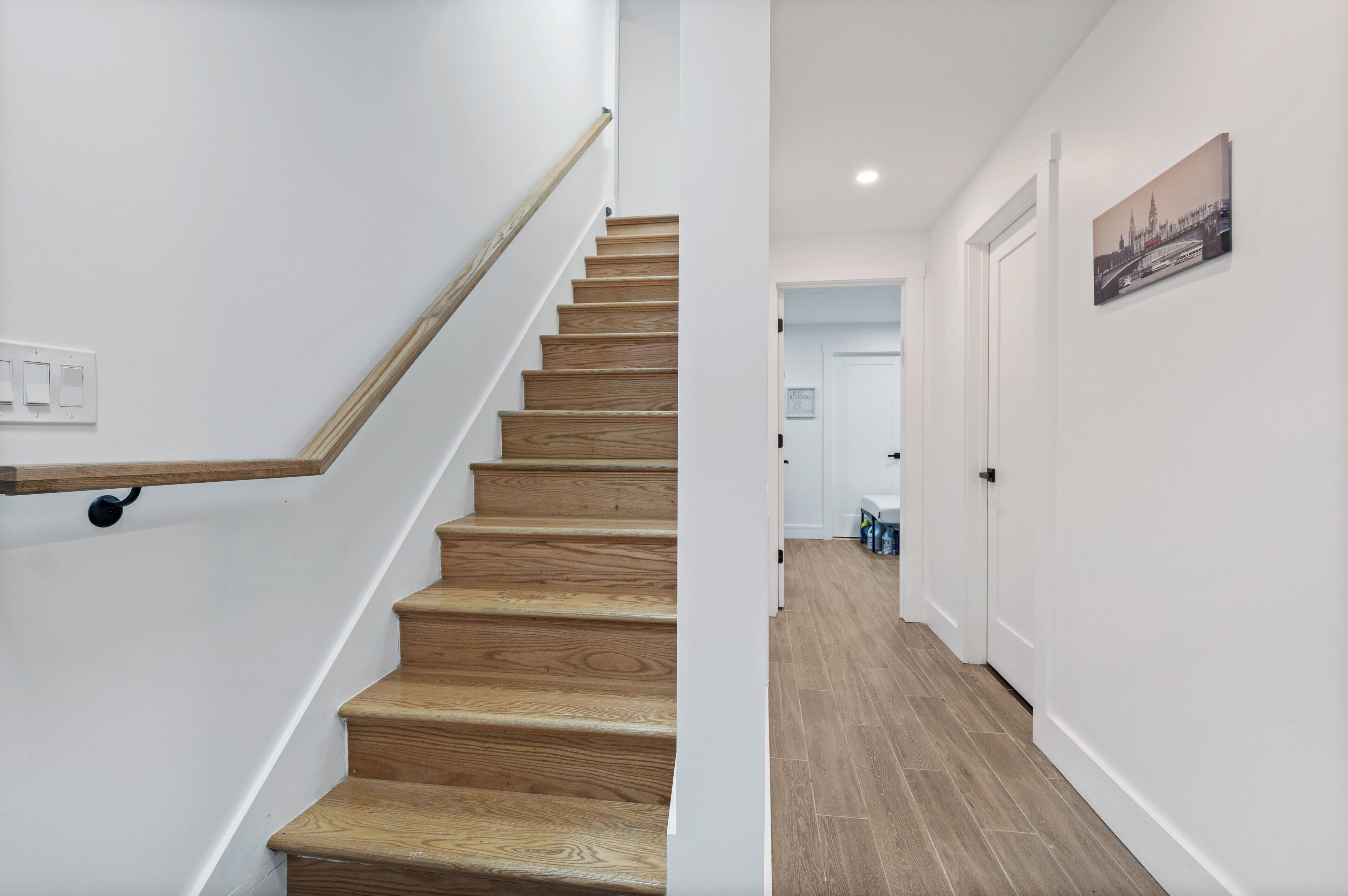 Una escalera de madera con barandilla, que lleva a un rellano con una puerta blanca y un cuadro enmarcado en la pared.