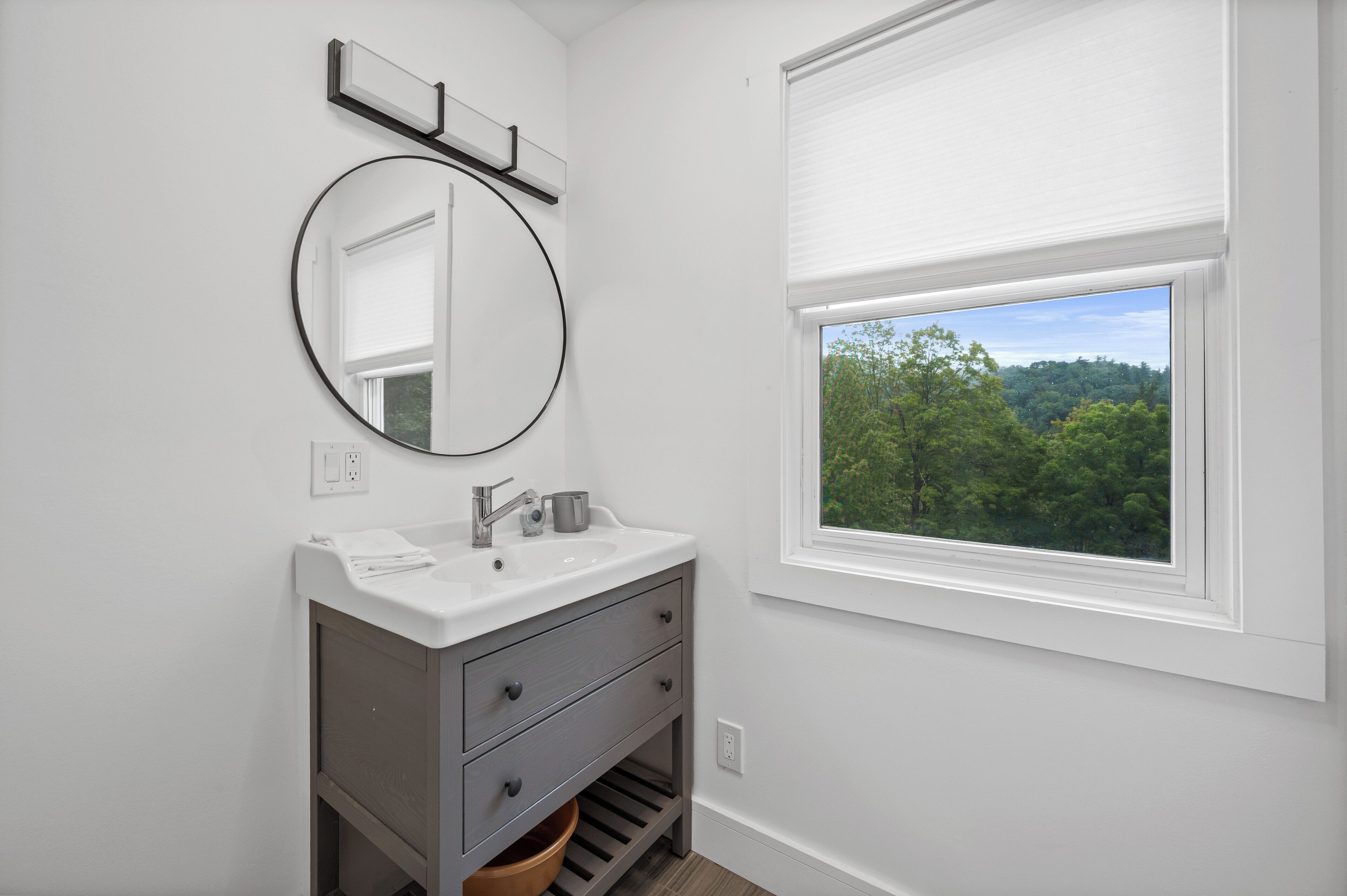 A bathroom with a white sink, a round mirror, and a window with a view of trees.