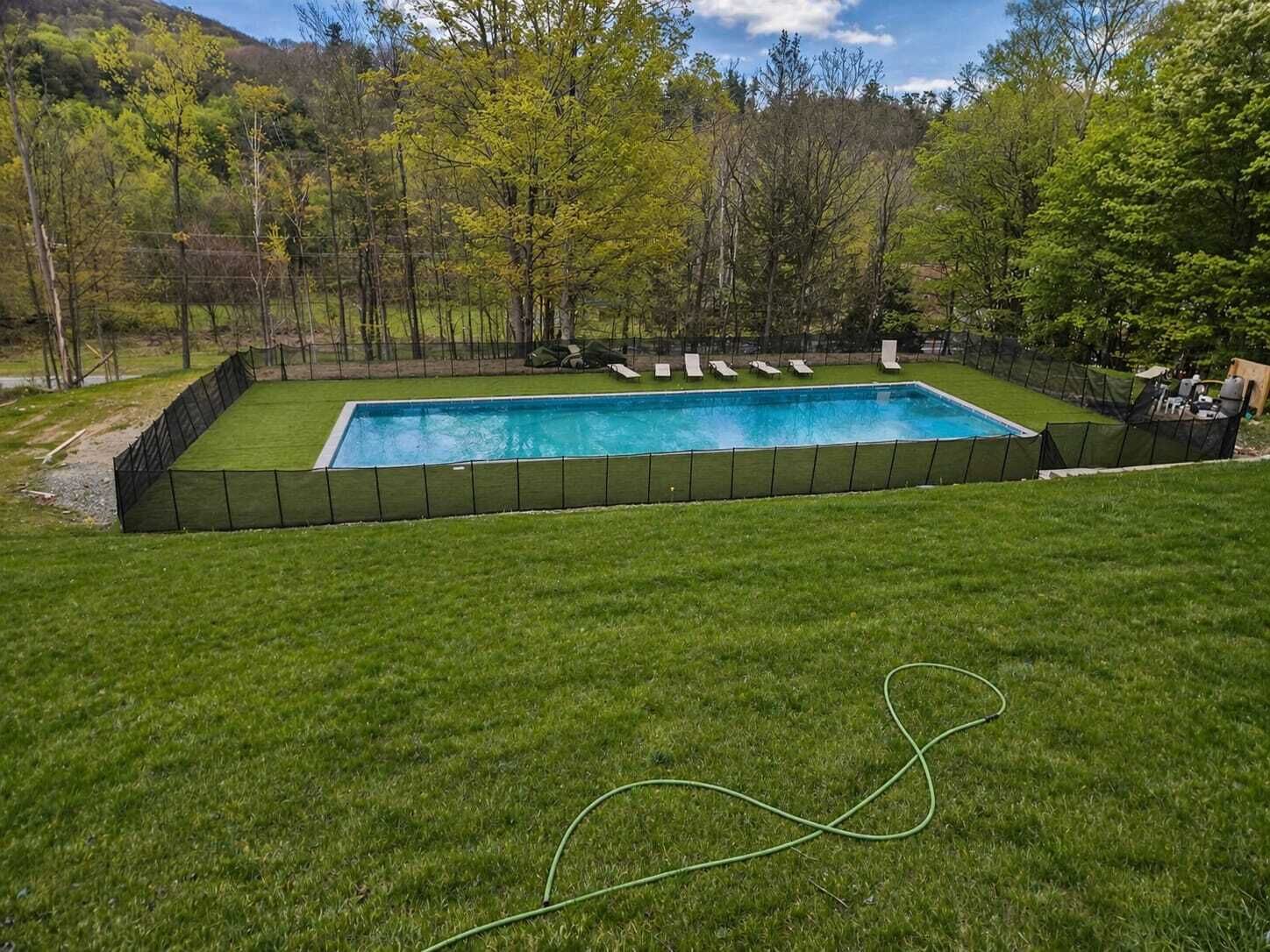 A rectangular swimming pool with a black fence, surrounded by a grassy lawn and trees.