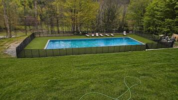 A rectangular swimming pool with a black fence, surrounded by a grassy lawn and trees.