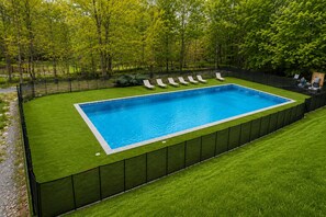 En rektangulær swimmingpool omgivet af et sort hegn, med en græsplæne og træer i baggrunden.