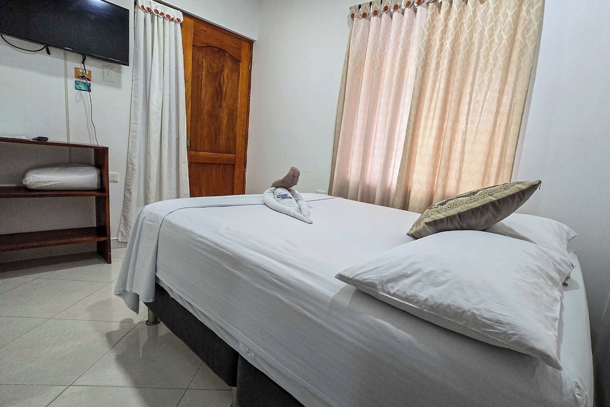 Un dormitorio con una cama, un armario de madera, una estantería con una toalla doblada y un televisor montado en la pared.