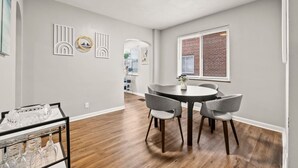 Un comedor con una mesa redonda y cuatro sillas, una mesita auxiliar con vasos, y una pared con paneles decorativos.