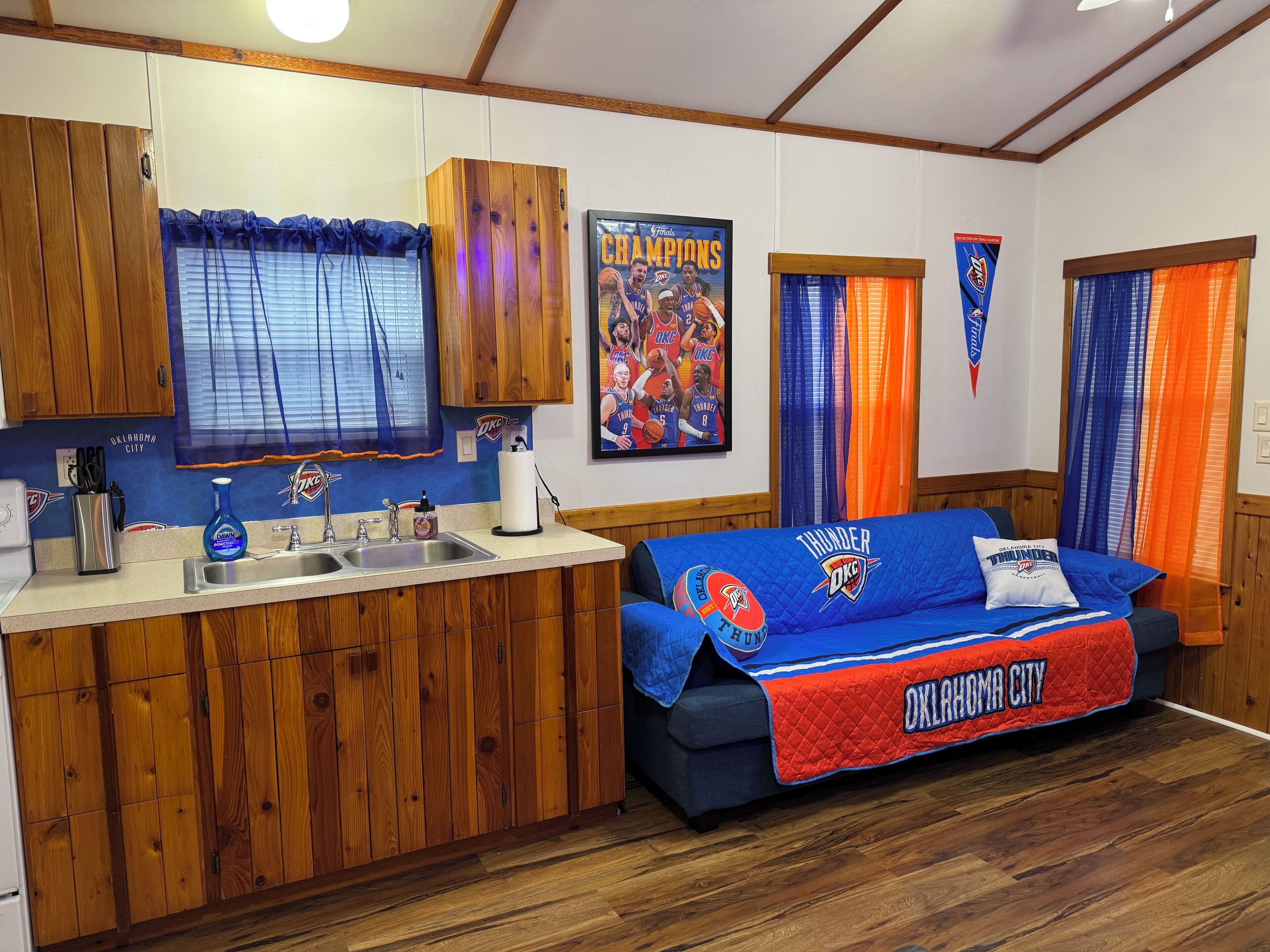 A room with a kitchenette, wooden cabinets, a blue couch with Oklahoma City Thunder branding, and sports-themed decorations.