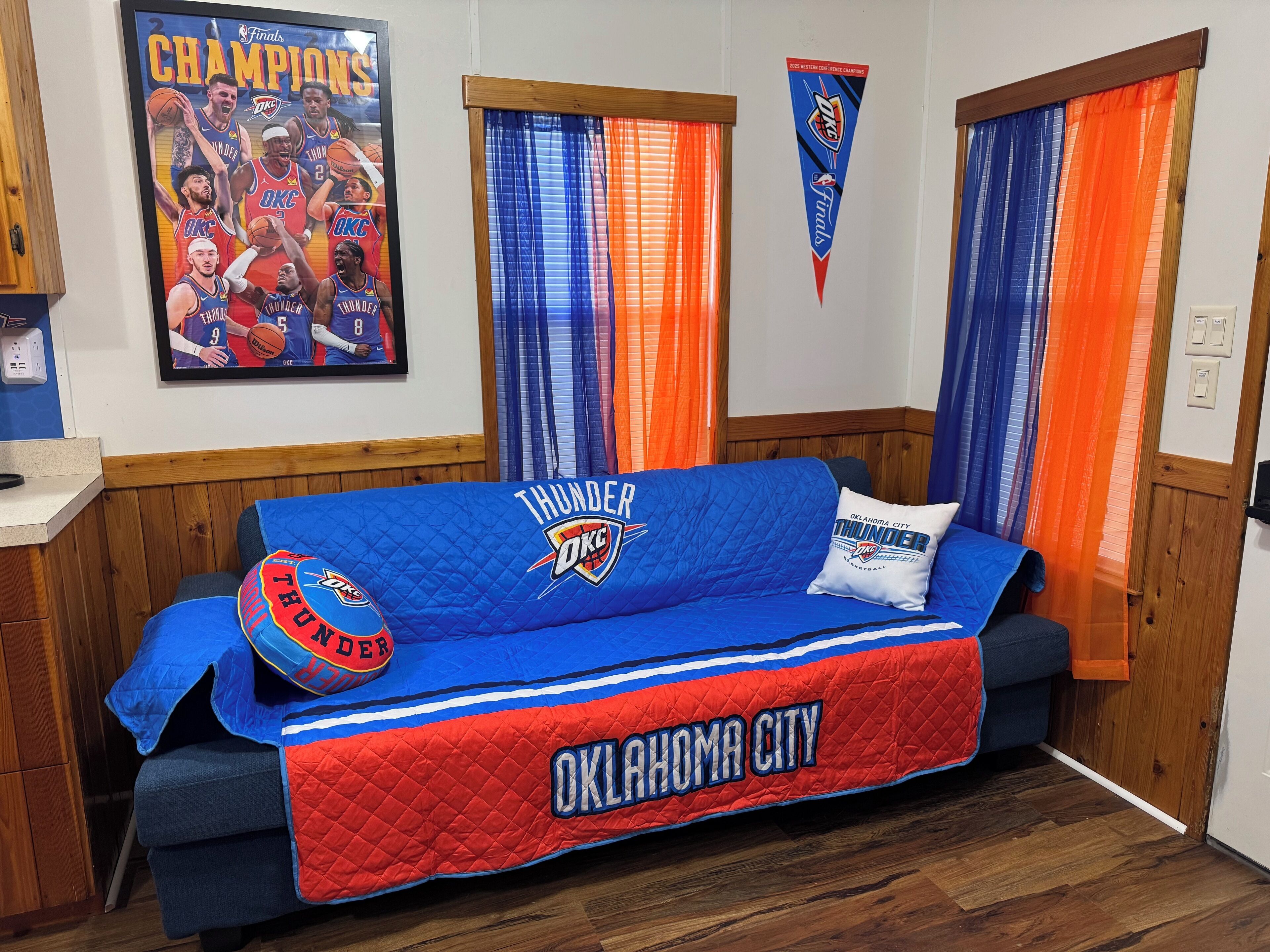 Ein Zimmer mit einem blauen und roten Sofa, das mit Oklahoma City Thunder Logopräsentationen, einem gerahmten Meisterschaftsposter und Vorhängen in Blau- und Orangefarben ausgestattet ist.