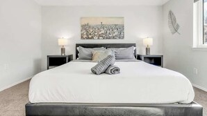 Un lit bien fait, avec une couette blanche et des serviettes pliées, deux lampes de chevet, une œuvre d’art représentant un champ accrochée au mur, et une fenêtre décorée de motifs végétaux.