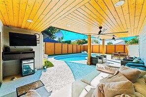 An outdoor living area with a pool, wooden ceiling, ceiling fans, and a seating area with cushions.
