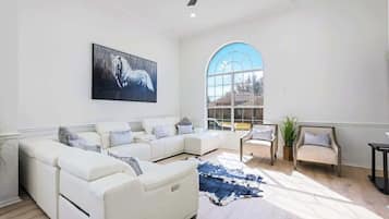Ein geräumiges Wohnzimmer mit einem weißen Modulsessel, einem großen Fenster und einem aufgehängten Bild eines Pferdes.