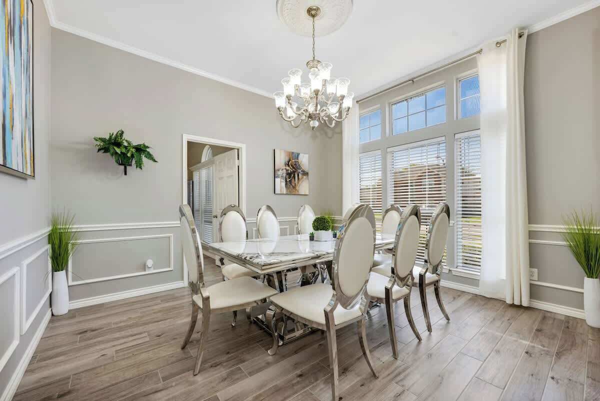 A dining room with a large table, white chairs, a chandelier, and large windows with blinds.