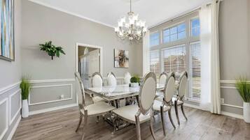 A dining room with a large table, white chairs, a chandelier, and large windows with blinds.