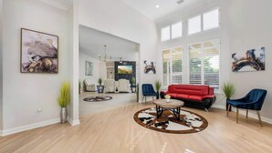 A spacious living room with a red sofa, blue chairs, and a round coffee table. There are large windows with blinds, artwork on the walls, and a ceiling light. - Exquisite 6 BR Retreat PoolSpa 4.5 Bath Sleeps 20+ (Richardson)