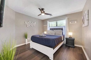 A bedroom with a bed, a nightstand, a ceiling fan, a window with blinds, and a wall-mounted sign. - Charming Oasis Hot Tub PingPong PS5 EV Sleeps 16+ (McKinney)