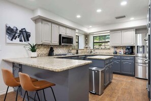 A modern kitchen with granite countertops, stainless steel appliances, and a central island with bar stools. - Charming Oasis Hot Tub PingPong PS5 EV Sleeps 16+ (McKinney)