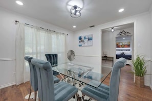A dining area with a glass table and chairs, a ceiling fan, and a painting on the wall. - Charming Oasis Hot Tub PingPong PS5 EV Sleeps 16+ (McKinney)