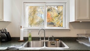 Una cuina amb un doble vàter d'acer inoxidable, una finestra que ofereix vistes a arbres d'autumn, un suport per ganivets i una ampolla de sabó per a plats.