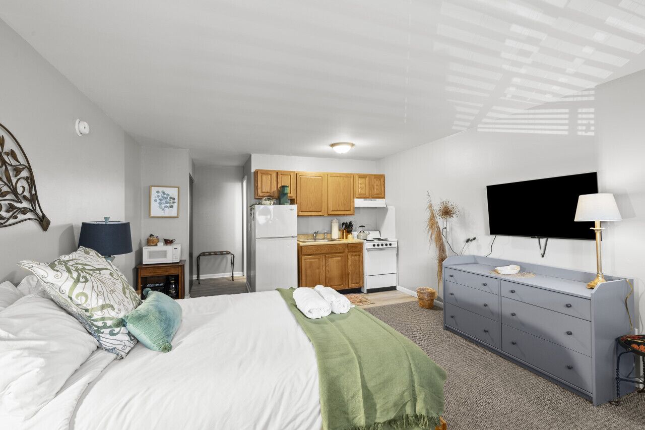 A modern bedroom with a bed, a dresser, a TV, and a kitchen in the background.