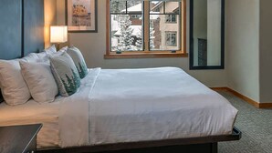 Una cama bien tendida con sábanas blancas y varios almohadones, una lámpara de noche y una ventana con vistas a un paisaje nevado.