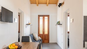 A dining area with a wooden table, chairs, and a fruit bowl. There is a television mounted on the wall and a door at the end of the room. - Costa Ovest Apartments (Nurachi)
