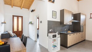 A modern kitchen with wooden cabinets, a microwave, and a stove. There is a dining area with a table and chairs, and a door leading to another room. - Costa Ovest Apartments (Nurachi)