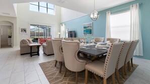 Un coin repas avec une table dressée pour six personnes, des chaises rembourrées de couleur beige, un lustre et une vue sur le jardin à travers de grandes fenêtres.