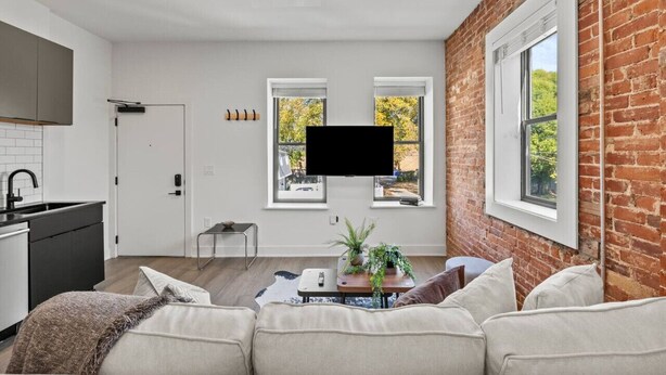 Een moderne woonkamer met een bakstenen muur, een flatscreen televisie boven een raam en een bank met kussens.