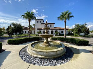 A circular fountain with water spouting from its center, surrounded by a stone border and pebbles. In the background, there is a white building with a brown roof and multiple windows, flanked by palm trees and well-maintained hedges. - Last Minute Disney Getaway! Villa with Private Pool & Hot Tub. Book Now! #18234 (Orlando)