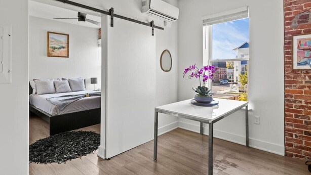 Ein modernes Schlafzimmer mit einem Bett, einem kleinen Tisch mit Blumenvase und Blick auf die Straße durch ein Fenster.