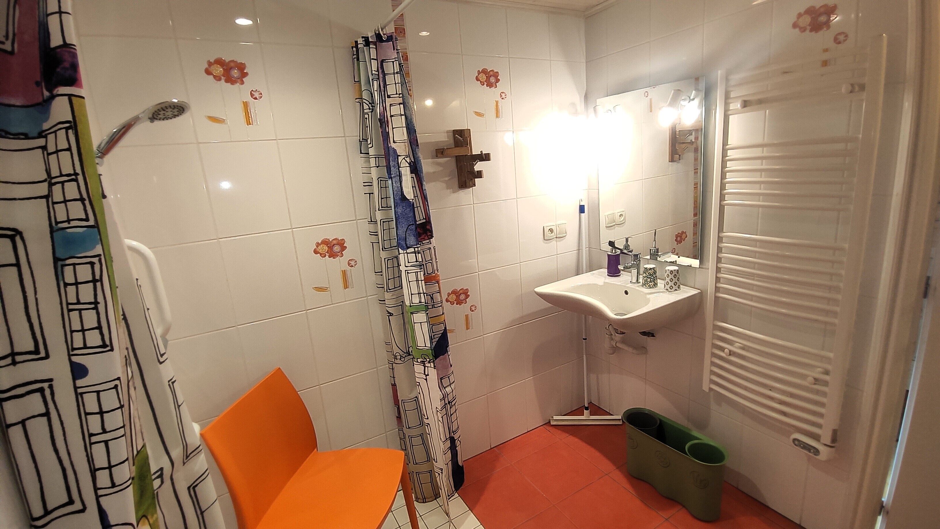 Een badkamer met een wit wastafel, spiegel en douchegordijn met bloemmotieven. Er staat een stoel en een afvalbak.