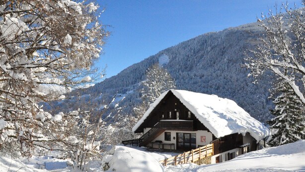 Un edificio de estilo chalet con techo cubierto de nieve, rodeado de árboles nevados y montañas al fondo.