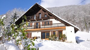 Un chalet de dos pisos con balcón de madera, rodeado de árboles cubiertos de nieve y un cielo despejado.