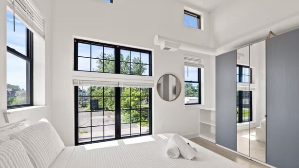 Un dormitorio moderno con una cama grande, un espejo redondo y vistas a zonas verdes por las ventanas.