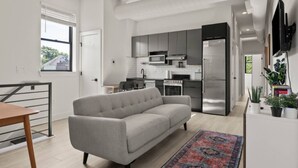 Un apartamento moderno con un sofá gris, una mesa de comedor, una cocina con electrodomésticos de acero inoxidable y un televisor montado en la pared.