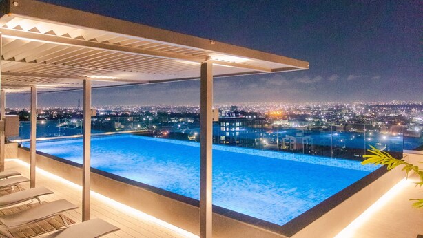 An infinity pool with a city view at night, a covered seating area with lounge chairs, and a well-lit poolside area. - Comfy One Bed Room Apartment at Lennox (Accra)