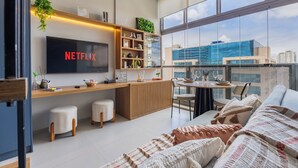 Ein modernes Wohnzimmer mit großem Fernseher, Holzmöbeln, einem Balkon mit Blick auf Gebäude und einer gemütlichen Sitzecke.
