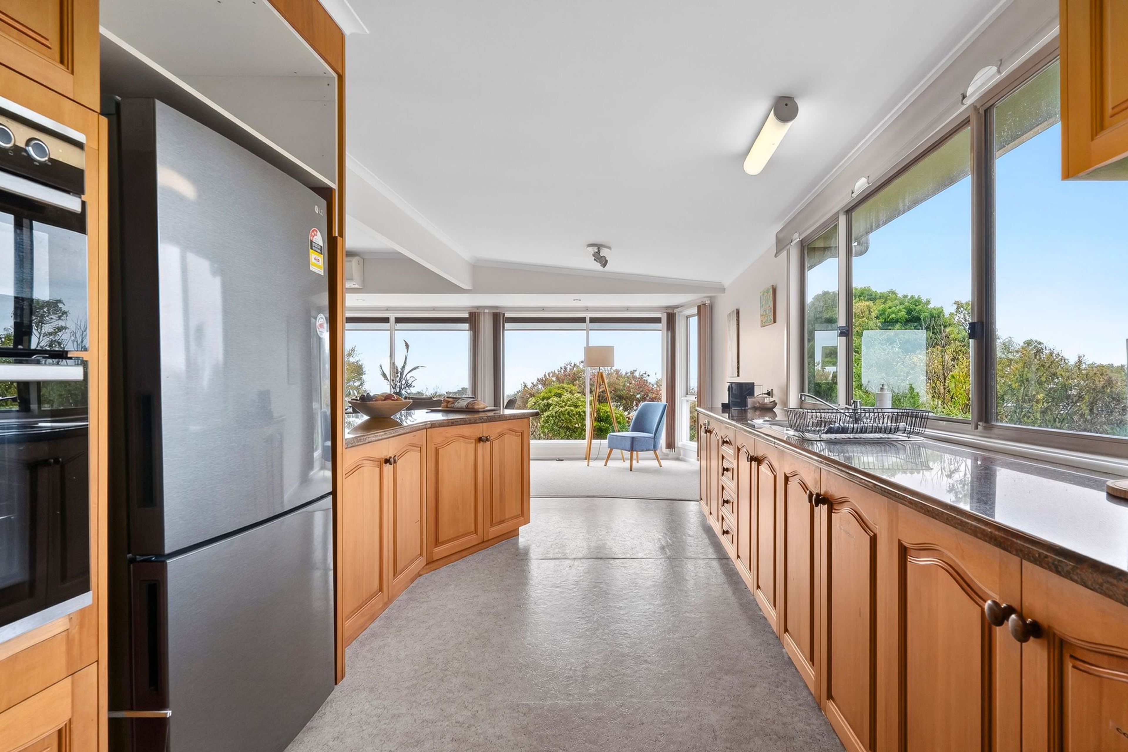 A modern kitchen with wooden cabinets, stainless steel appliances, and large windows offering a view of greenery.
