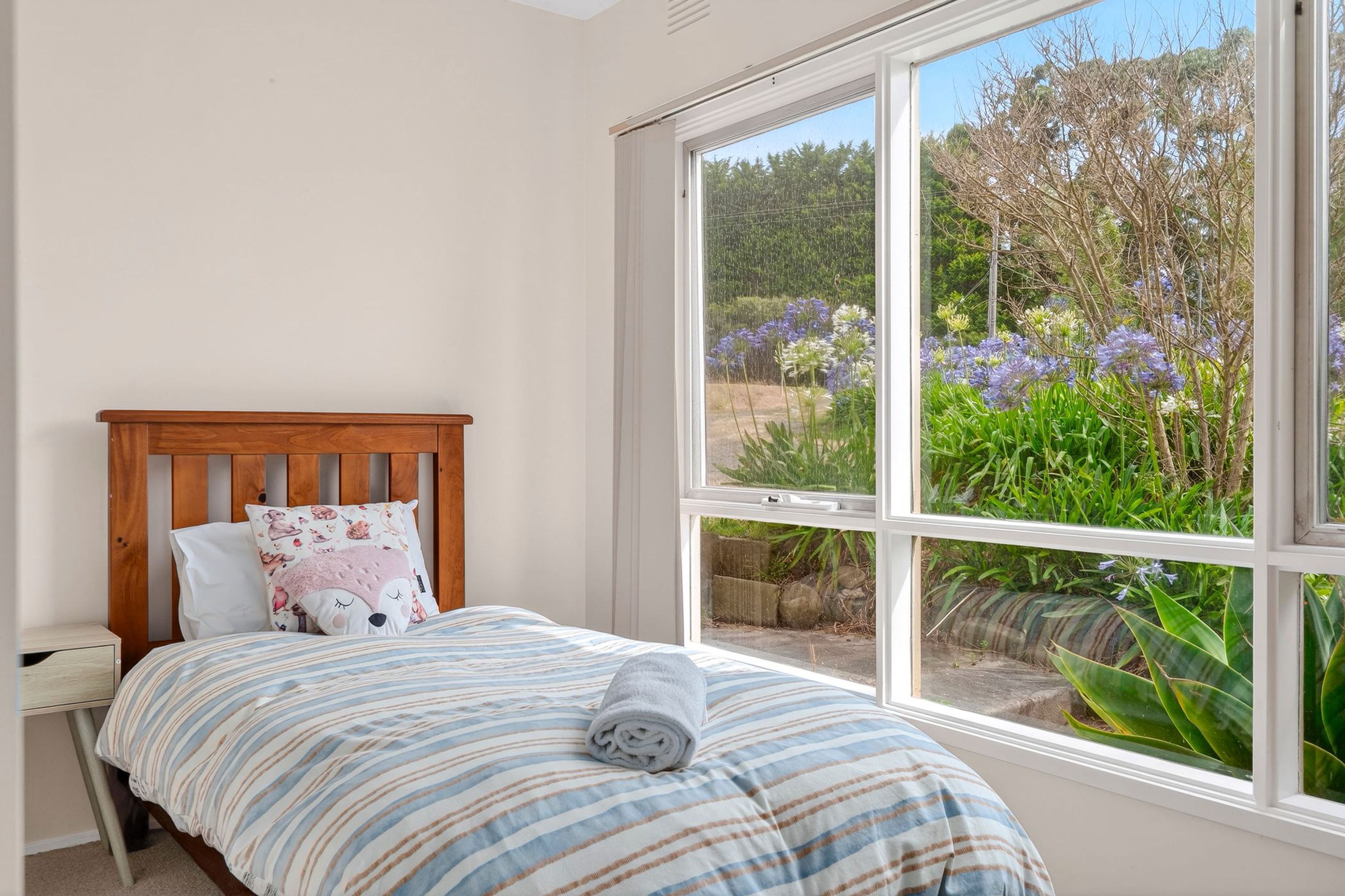 A bedroom with a wooden headboard, a bed with striped bedding, a pillow with a dog design, and a view of a garden through a large window.